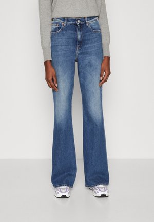 Person wearing high-waisted blue flare jeans with faded front thighs, gray long-sleeve top, and white and purple Nike sneakers.