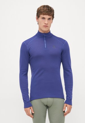Junger Mann, der ein eng geschnittenes, langärmeliges, blaues Zip-Up-Oberteil und eine hellgraue Hose trägt, steht vor einem schlichten weißen Hintergrund.