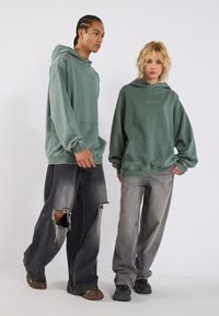 Felpe verdi con cappuccio e tasche a marsupio, vestibilità rilassata. Indossate con jeans neri e grigi strappati. Entrambi indossano sneaker. Dettaglio ricamato semplice.