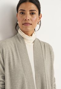 Vrouw met donker haar naar achteren, met zilveren creolen, een crèmekleurige coltrui en een lichtgrijze cardigan, tegen een plain witte achtergrond.