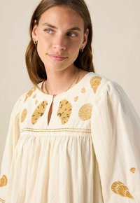 Weiße Bluse mit goldener Muschelstickerei, runder Halslinie, Schlüssellochöffnung und voluminösen Ärmeln. Leichter Stoff und strukturierte Details.