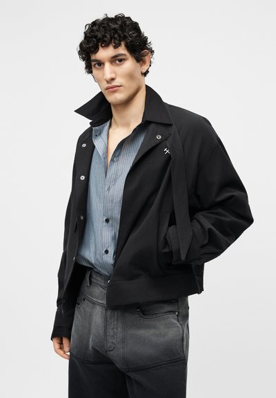 Junger Mann mit lockigem schwarzem Haar trägt eine schwarze Jacke über einem blau gestreiften Hemd und verblassten schwarzen Hosen und steht vor einem einfarbigen Hintergrund.