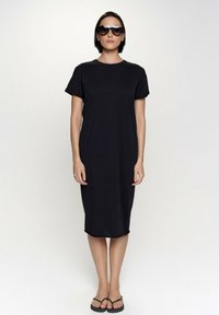 Czarna sukienka do kolan wykonana z lekkiego materiału, z krótkimi rękawami i okrągłym dekoltem. Noszona z ciemnymi okularami przeciwsłonecznymi i klapkami.