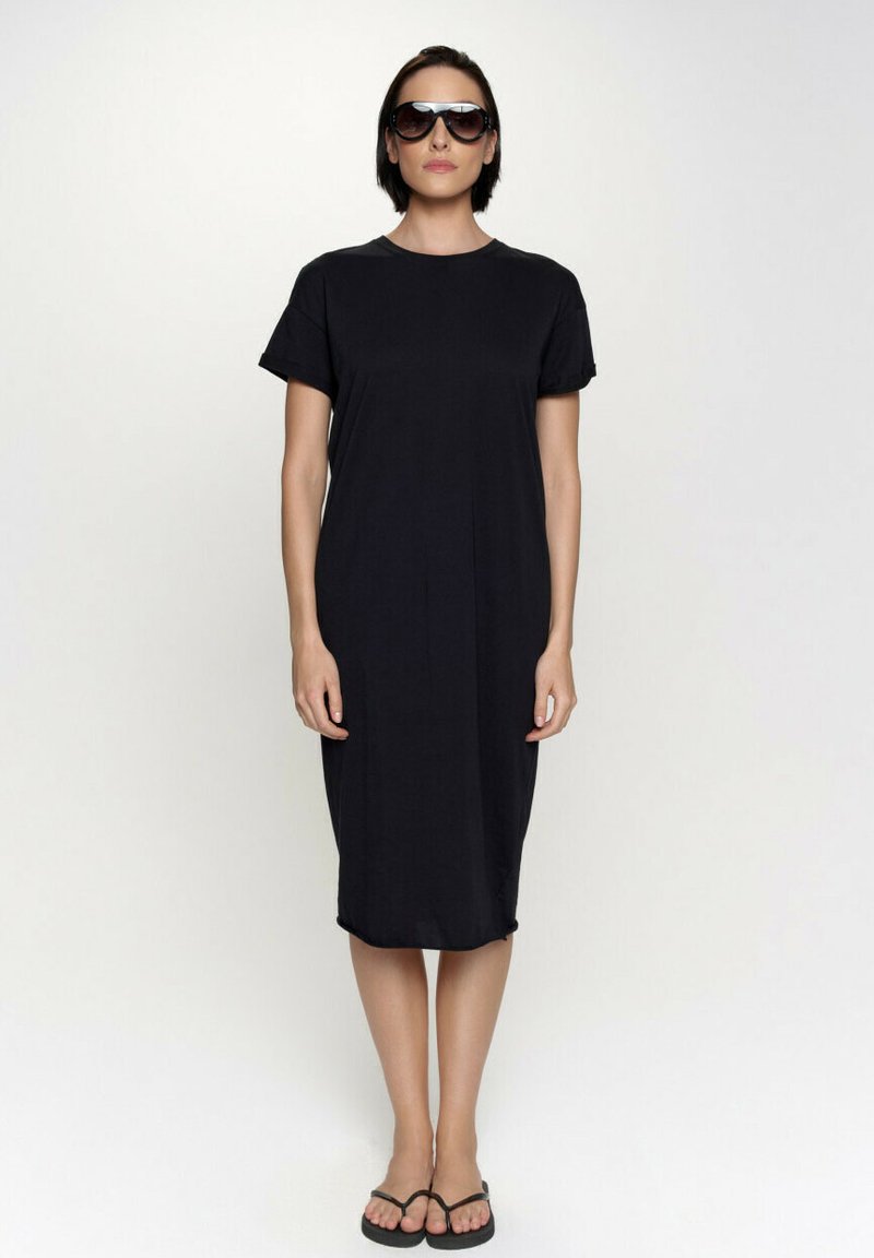 Czarna sukienka do kolan wykonana z lekkiego materiału, z krótkimi rękawami i okrągłym dekoltem. Noszona z ciemnymi okularami przeciwsłonecznymi i klapkami.