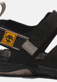 Svart sandal med texturerade remmar, en grå accentpanel och en gul logotypklistermärke. Designad med en stödjande sula för stabilitet.