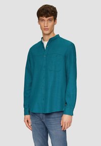Teal overhemd met een Chinese kraag, borstzak en een structuurstof. Gecombineerd met blauwe jeans staat het model tegen een grijze achtergrond.