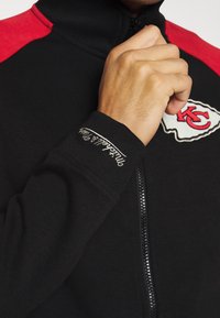 Nahaufnahme einer Hand, die eine schwarz-rote Jacke mit dem Logo der Kansas City Chiefs und weiß gesticktem Text am Ärmel zippt.