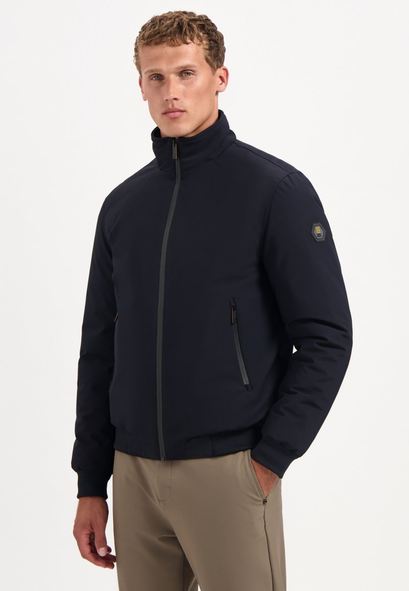 Marine bombehanger met een hoge kraag, rits aan de voorkant en twee zijzakken. Heeft een subtiele textuur en een hexagonaal logo op de mouw.