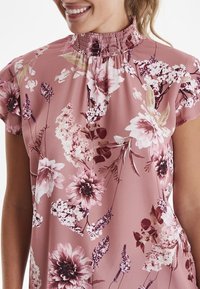 Blusa floreale in morbido tessuto rosa chiaro, con un alto colletto smock, maniche corte e vari motivi floreali in crema e bordeaux.