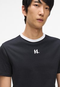 Černé tričko s bílým kulatým límcem a krátkými rukávy. Na hrudi se nachází malé bílé vyšívané logo. Jednoduchý, obepínající design.