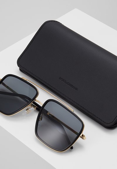Gafas de sol cuadradas en negro y dorado con lentes oscuras junto a un estuche de cuero negro con la marca Dolce & Gabbana sobre una superficie blanca.