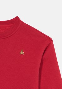 Κόκκινο φούτερ με στρογγυλή λαιμόκοψη και μικρό κεντημένο καφέ λογότυπο αρκουδάκι στο πάνω αριστερό μέρος του στήθους, απλό φόντο.