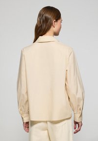 Camicia in cotone beige con colletto, vestibilità comoda e maniche lunghe arricciate. Presenta una texture liscia e polsini classici con bottoni.
