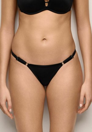 Femme portant un bikini noir avec des bretelles latérales ajustables, montrée du ventre jusqu'aux cuisses sur un fond uni.