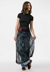 T-shirt nera aderente, gonna lunga in pizzo e pantaloni di denim sovrapposti. Borsa a cintura bordeaux con un accento a chiusura. Scarpe scure completano l'outfit.