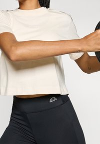 Osoba w beżowej krótkiej koszulce i czarnych legginsach, trzymająca w obu rękach ciężar do podnoszenia, z widocznymi tatuażami na ramionach.