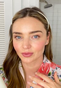 Mannequin avec de longs cheveux ondulés, portant un peignoir rose à motifs floraux et un bandeau en perles, avec un maquillage naturel et une bouteille à la main.