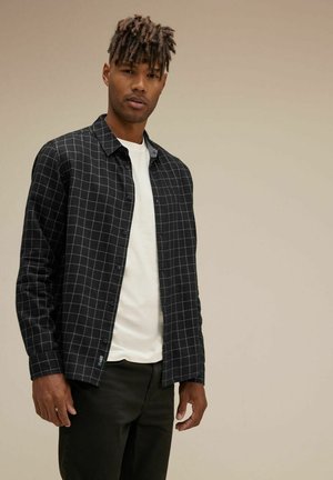 Camisa de cuadros blanco y negro de botones sobre una camiseta blanca, exhibida con pantalones negros. Tejido suave y patrón de cuadrícula clara.