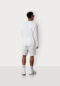 Weißes Sweatshirt mit langen Ärmeln, kombiniert mit passenden weißen Shorts. Das Outfit zeichnet sich durch eine weiche Textur und einen lässigen Schnitt aus. Weiße Turnschuhe vervollständigen den Look.