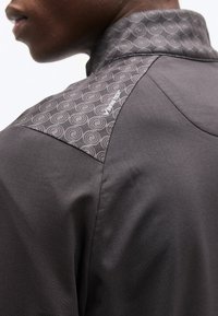 Κοντινό πλάνο ενός ατόμου που φοράει σκούρο μπουφάν με μοτίβο στον ώμο, που περιλαμβάνει σπειροειδή σχέδια και ένα μικρό λευκό λογότυπο.