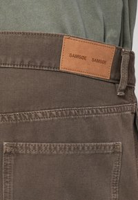 Pantalones de tela marrón con un parche de cuero en la cinturilla que dice "SAMSNØ". Presenta un corte clásico y un detalle de costura en el bolsillo trasero.
