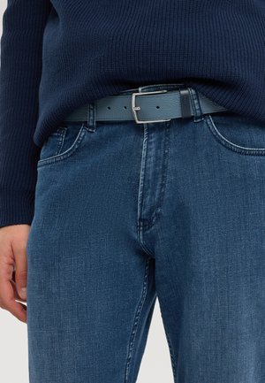 Close-up van een persoon die een blauwe denim spijkerbroek draagt met een bijpassende blauwe leren riem en een marineblauwe gebreide trui.