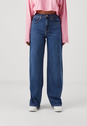 Persoon draagt een high-waisted wijde blauwe jeans, roze cropped top met lange mouwen en witte sneakers, staand op een neutrale achtergrond.