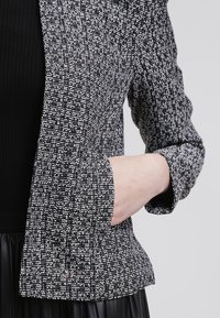 Personne portant une veste texturée noire et blanche avec une main placée dans la poche avant sur un fond uni.