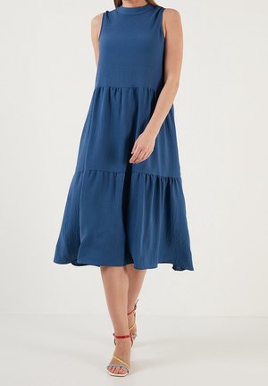 Niebieska sukienka midi bez rękawów z falbanami, noszona z wielokolorowymi sandałami na wysokim obcasie, modelka idzie na tle jednolitego jasnego tła.