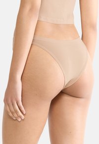 Nude-kleurige naadloze bikini slip met een hoge uitsnijding, zachte textuur en een minimale tailleband, die een close-up weergave van de pasvorm aan de achterkant toont.