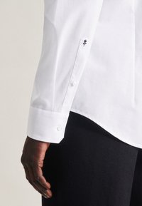 Weißes Hemd mit langen Ärmeln und knöpfbaren Manschetten. Die Manschette ist mit einem kleinen schwarzen Stickmuster versehen. Glatter Stoff, maßgeschneiderte Passform, kontrastierende Knöpfe.