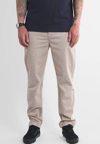 Pantalon en coton beige à coupe droite, doté de poches latérales et de détails de couture subtils. Associé à des baskets noires texturées.