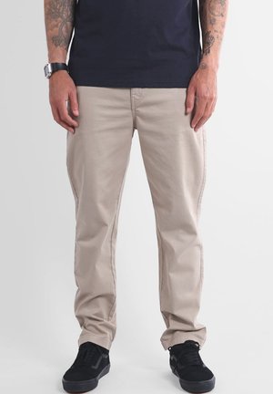 Pantalon en coton beige à coupe droite, doté de poches latérales et de détails de couture subtils. Associé à des baskets noires texturées.