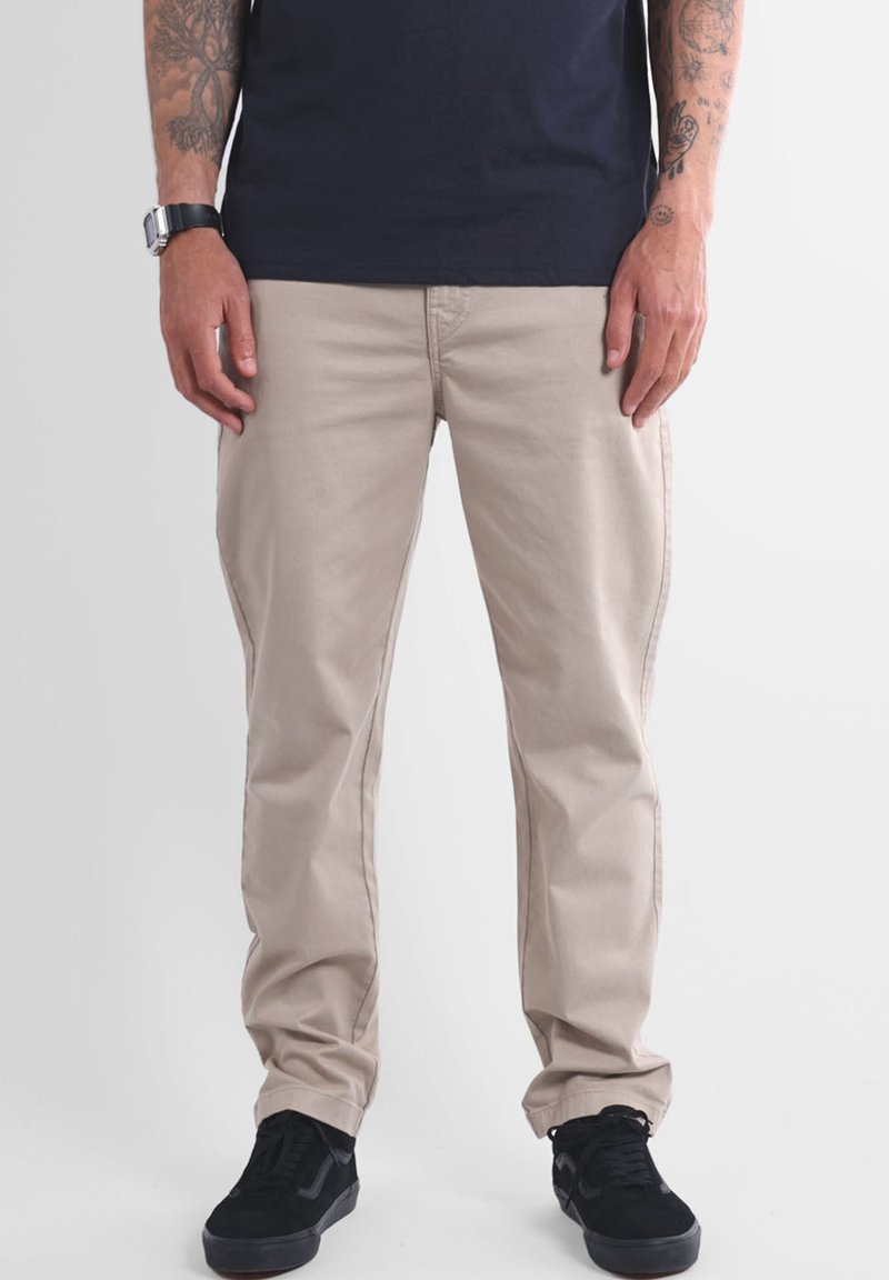 Pantalon en coton beige à coupe droite, doté de poches latérales et de détails de couture subtils. Associé à des baskets noires texturées.