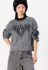 Grijze gebreide trui met een pluizige textuur, ronde zwarte halslijn en abstract donker patroon aan de voorkant. Gecombineerd met grijze broek.