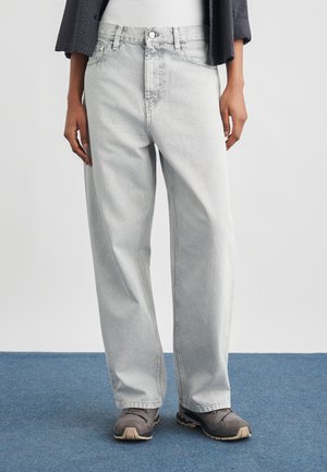 Άτομο που φορά ανοιχτό γκρι τζιν άνετης εφαρμογής και γκρι αθλητικά παπούτσια, στέκεται σε μπλε χαλί μπροστά σε λευκό τοίχο.