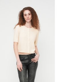 Νεαρή γυναίκα με σγουρά κόκκινα μαλλιά, φορώντας μπεζ κοντομάνικο πλεκτό ζακετάκι με κουμπιά και μαύρο υφασμάτινο παντελόνι, στέκεται μπροστά σε λευκό απλό φόντο.