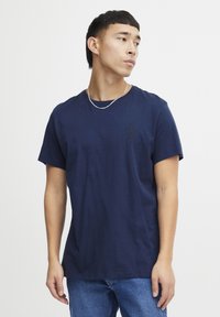 Temno modra bombažna majica s kratkimi rokavi, okroglo izrezana in z majhnim črnim logotipom na levem prsnem predelu. Gladka textura, sproščen kroj.