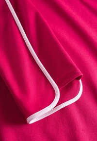 Fuchsiafarbenes Textil mit glatter Oberfläche und weißen Paspeln an den Kanten. Zeigt ein gefaltetes Ärmel-Detail auf einer flachen Oberfläche.