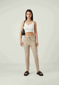 Top crop blanc avec des bords en scallop, associé à un jogging beige rayé et des sandales noires à enfiler. Le mannequin se tient devant un fond neutre.