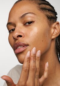 Gros plan sur le visage d'une personne avec une peau lisse et brillante. Le sujet a des cheveux tressés, des lèvres d'un brun clair et des ongles naturels. Des tatouages visibles sur la main.