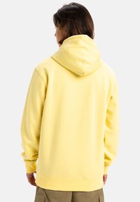 Sudadera amarilla con una gran capucha de tela suave. Presenta puños y dobladillo acanalados, un ajuste extragrande y una textura lisa. Diseño minimalista.