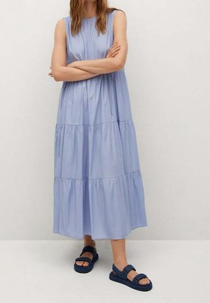 Frau trägt ein ärmelloses hellblaues Stufen-Midikleid mit dunkelblauen Sandalen und steht mit verschränkten Armen vor einer schlichten hellen Wand.