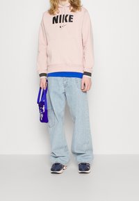 Světle růžová Nike mikina s černým logem, modrá vrstvená košile, volné světle modré džíny a námořnické tenisky. Drží fialovou tašku s bílým designem.