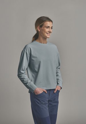 Smiling woman with hands in pockets wearing a light blue long-sleeve shirt and dark blue pants, standing against a plain gray background.