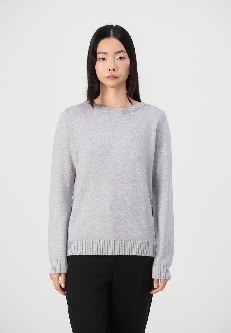 Helles grauer Strickpullover mit Rundhalsausschnitt, gerippten Bündchen und Saum. Weiche Textur, lange Ärmel und lässige Passform. Kombiniert mit schwarzen Hosen.