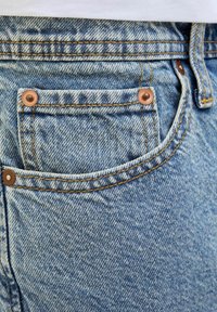 Light blue denim jeans with a visible pocket, copper rivets, contrasting yellow stitching, and a textured fabric finish.