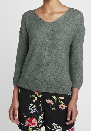 Zielony sweter dzianinowy o fakturze z rękawami do łokci, zestawiony z czarnymi szortami zdobionymi kolorowym kwiatowym wzorem.