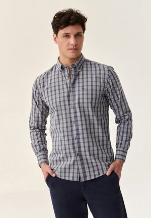 Button-up shirt in navy and white plaid pattern, long sleeves, standard collar, with a subtle logo detail on the chest. Smooth texture.