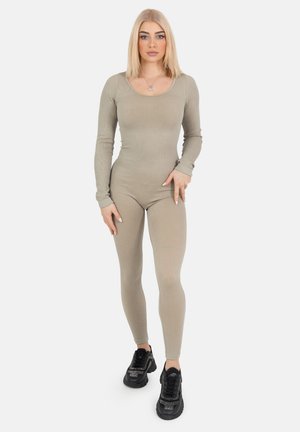 Ribbet, langærmet jumpsuit i lys beige, tætsiddende design, parret med sorte sneakers med glitrende accenter. Enkel rund halsudskæring, ingen mønstre.